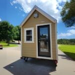 Tan Tumbleweed Farallon tiny house on a dual-axle trailer: steep silver gable, white trim, black-framed glass door, parked on pavement under a blue sky