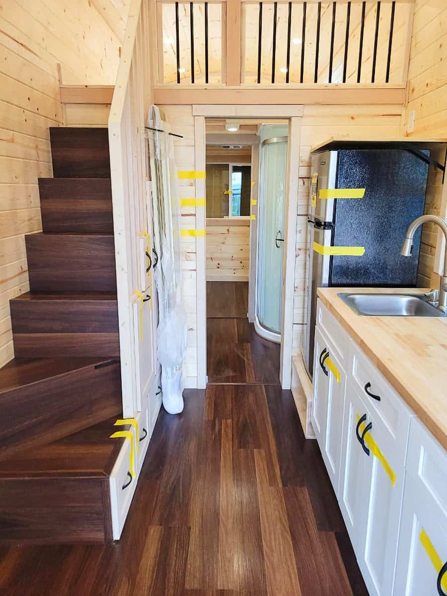 Interior looking down the hall: dark storage stairs to a loft on the left, white kitchen and fridge on the right, curved glass shower beyond