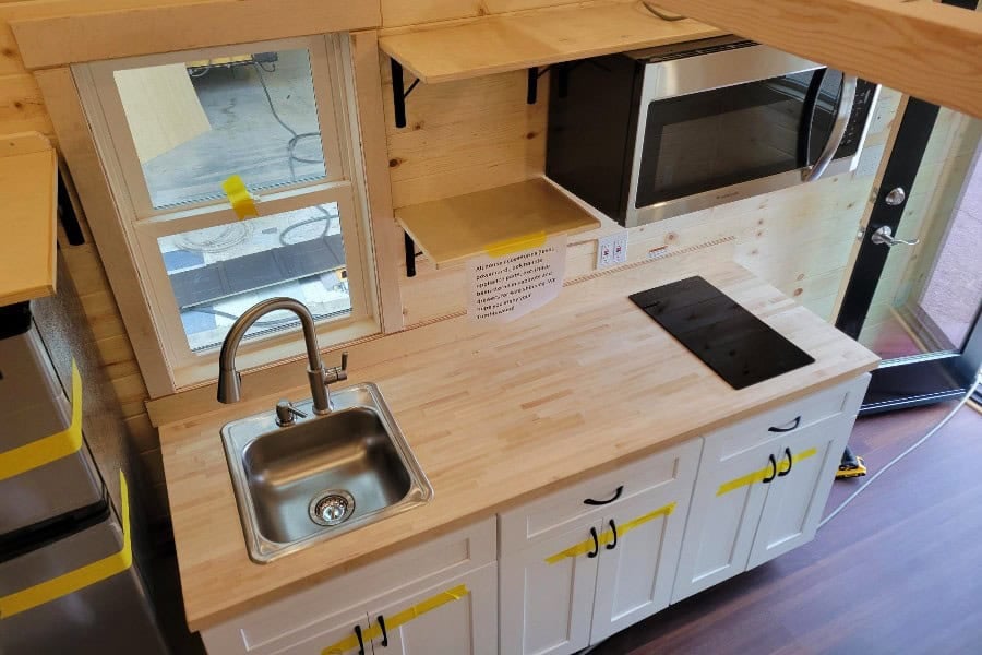 Maple-counter kitchen with stainless sink, two-burner cooktop, microwave on a wood shelf, white cabinets with yellow tape still on the pulls