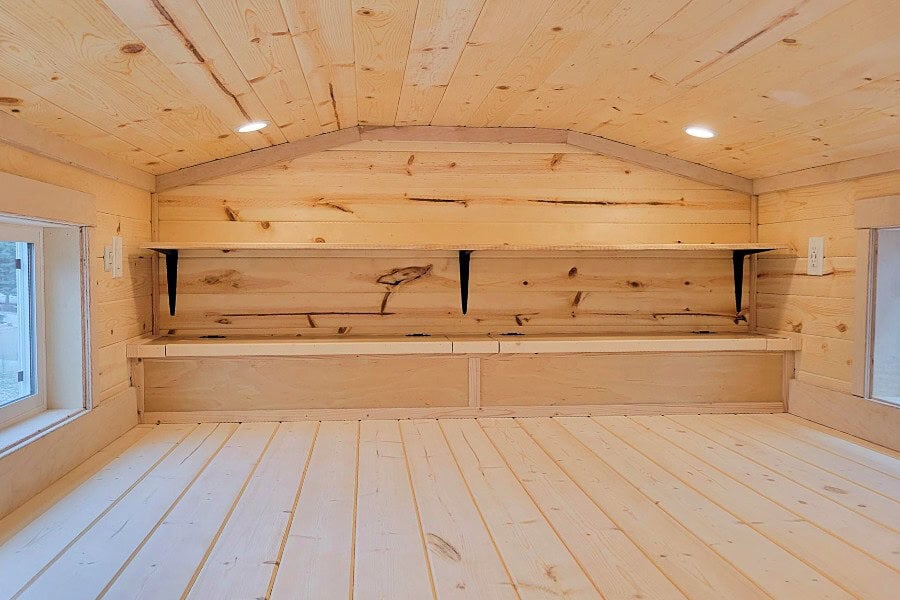 Pine sleeping loft under the gable: two small side windows, built-in bench at the far wall, long shelf on black brackets, two recessed lights