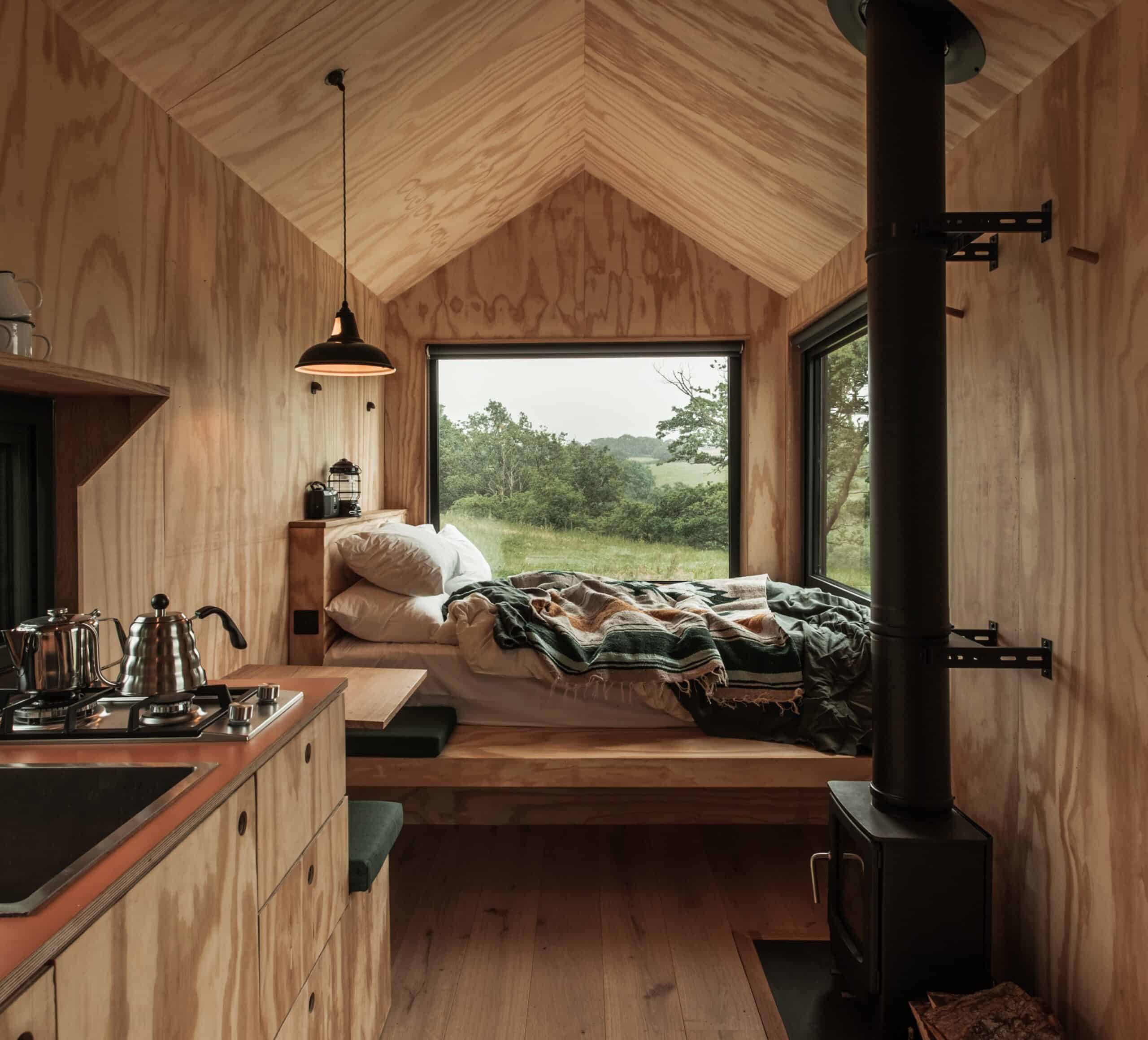 Cabin interior looking toward the bed under a large end window, wood-burner and black flue on the right, kitchen counter and gas stove in the foreground