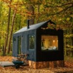 Dark gabled cabin on log piers in autumn leaves, black vertical siding, chimney on the ridge, two chairs and a table on a low deck, teal wheelbarrow in the leaves, bed visible through the end window
