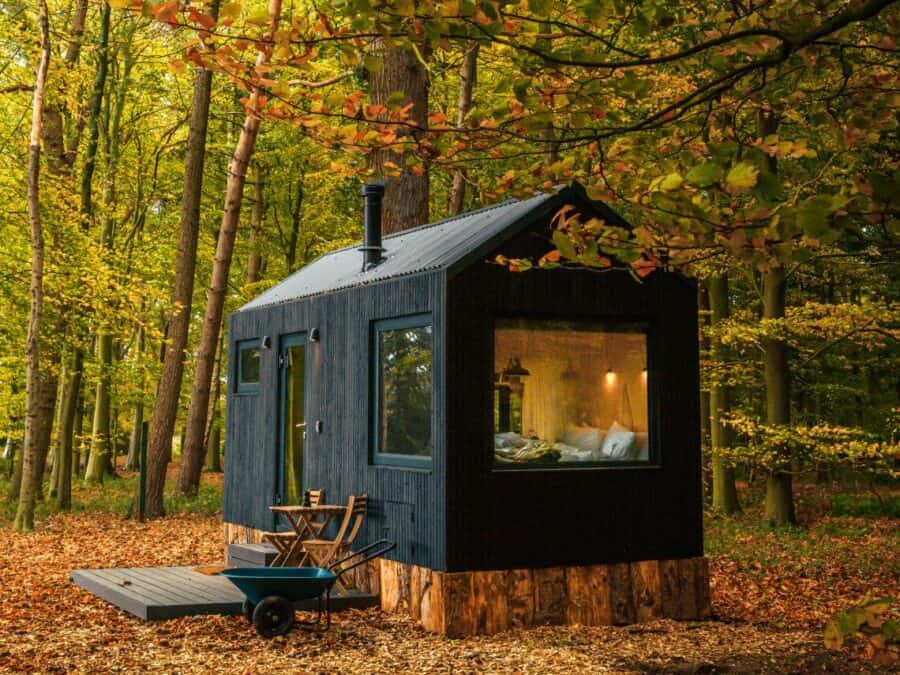 Dark gabled cabin on log piers in autumn leaves, black vertical siding, chimney on the ridge, two chairs and a table on a low deck, teal wheelbarrow in the leaves, bed visible through the end window