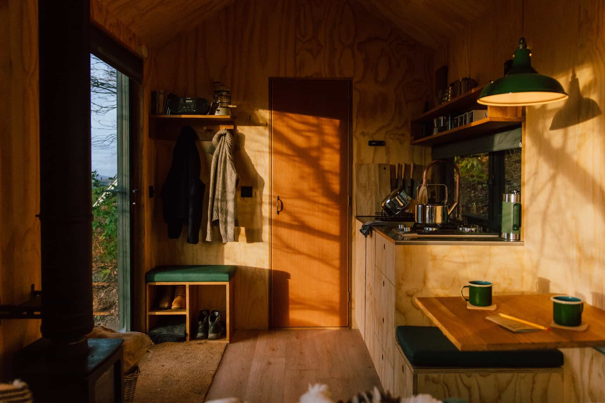 Plywood cabin living run: wood-burning heater at left, built-in shoe bench with green cushion and coats on pegs, kitchen with gas stove, small table with two green mugs under a green pendant