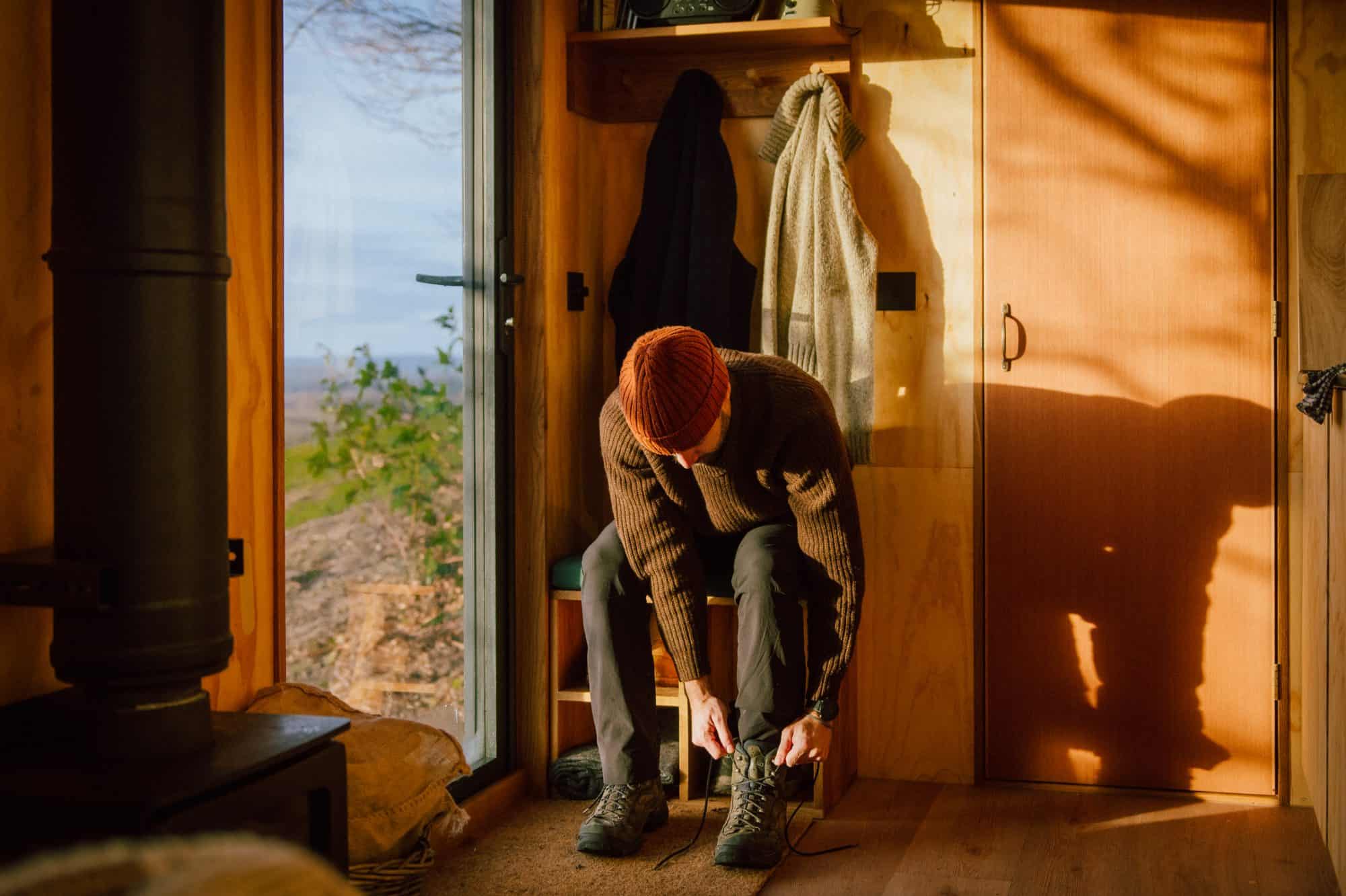 Person in an orange beanie tying hiking boots on a built-in plywood shoe bench, coats on pegs, wood-burner pipe at left, glass door to the hills