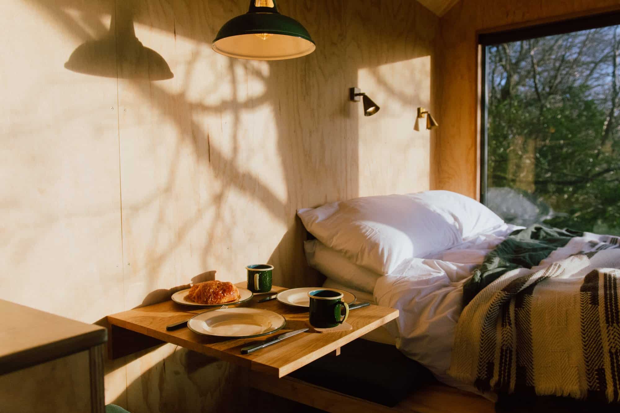 Plywood cabin interior: white bedding with a green throw and patterned blanket, two reading lights, a window into the trees, a small square table set for two with plates and green mugs