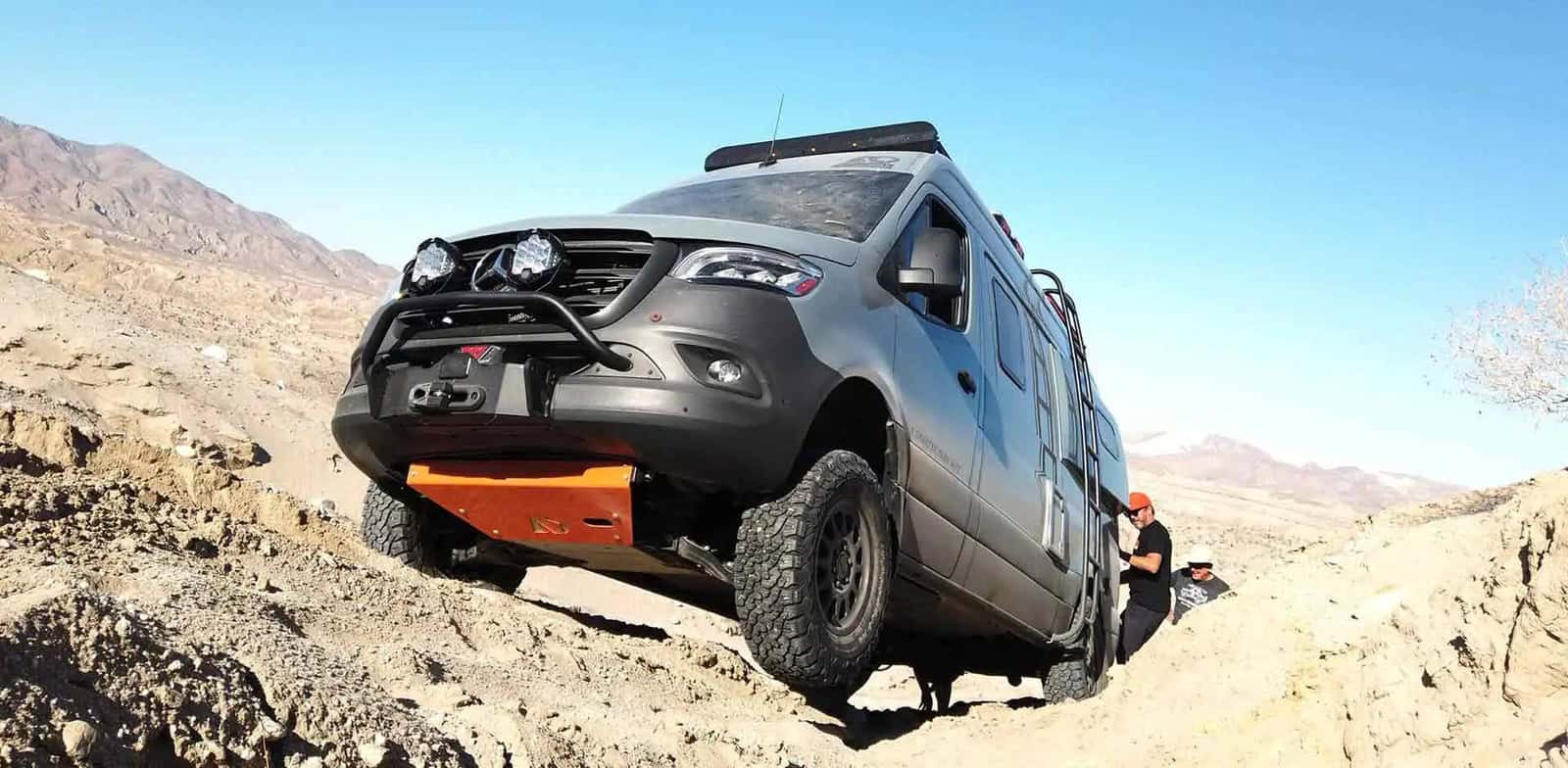 Grey 4x4 Sprinter climbing a rocky dirt incline, two people spotting from the ground