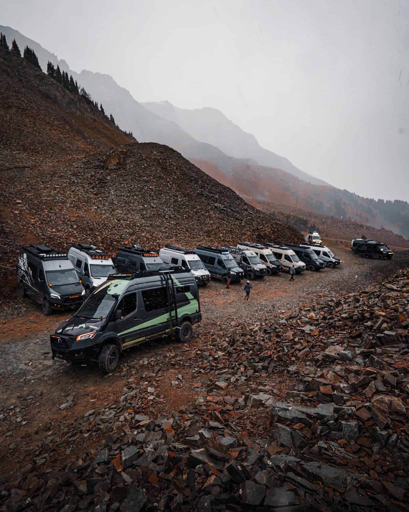 Line of 4x4 adventure vans parked on a rocky Colorado mountain slope under low cloud