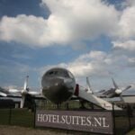 Head-on exterior of a four-engine propeller airplane on grass at Teuge, boarding stairs to the fuselage door, hotelsuites.nl banner on the fence