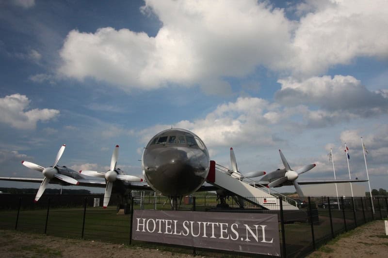 Head-on exterior of a four-engine propeller airplane on grass at Teuge, boarding stairs to the fuselage door, hotelsuites.nl banner on the fence