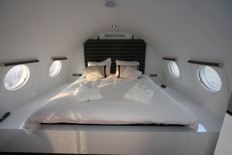 Kingsize bed with white linens at the tail, four round porthole windows, folded robes