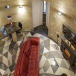 High-angle view of a circular loft living room: tufted red leather sofa facing a black electric fireplace, geometric tile floor, porthole windows, and a silver spiral stair