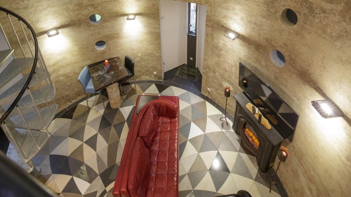 High-angle view of a circular loft living room: tufted red leather sofa facing a black electric fireplace, geometric tile floor, porthole windows, and a silver spiral stair