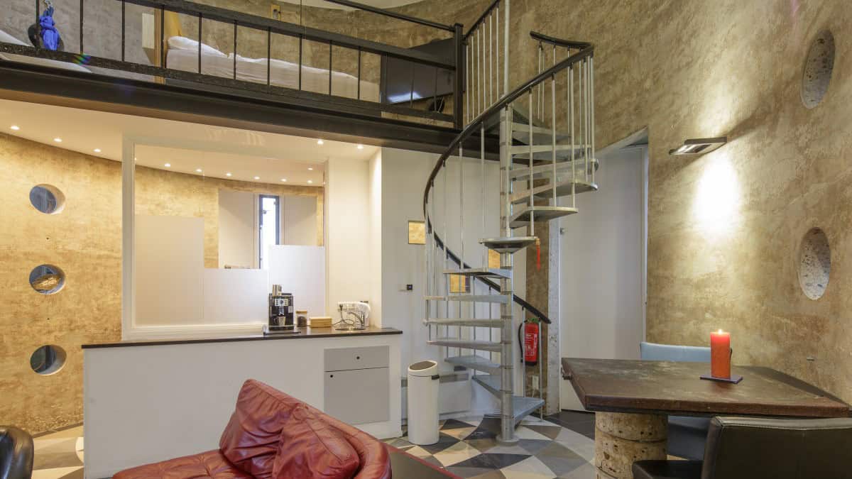 Circular loft looking toward a galvanized spiral stair and a mezzanine queen with white linens, kitchenette behind a white counter, red sofa in the foreground