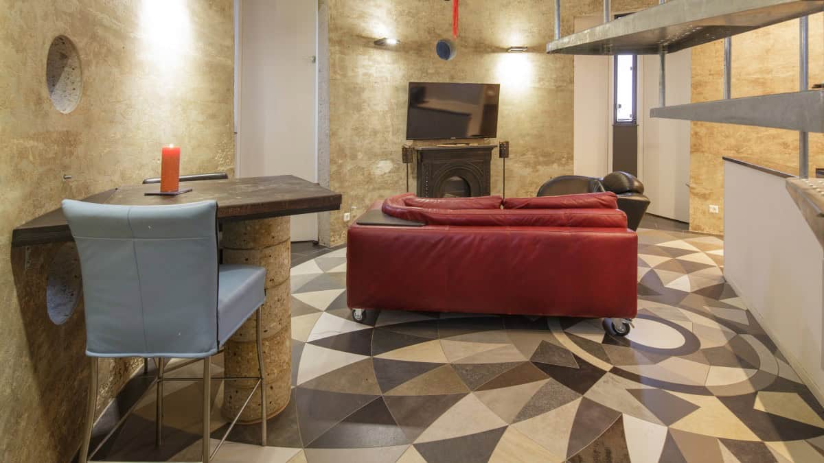 Eye-level circular living room with a red leather sofa on casters facing a black fireplace and wall TV, geometric radiating tile, and two porthole windows