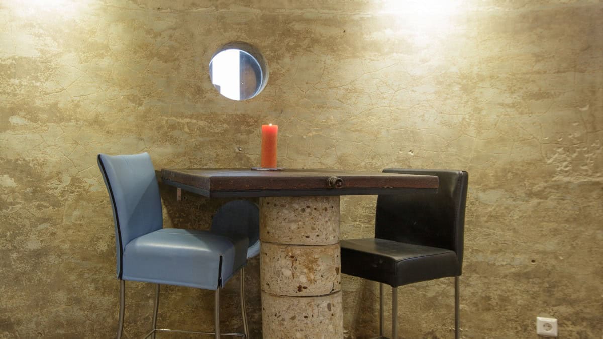 Small square table on stacked concrete cylinders against a curved textured wall, orange pillar candle, one blue chair and one black chair, porthole above