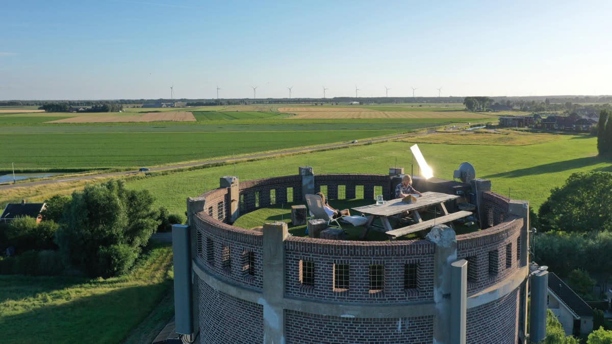 Aerial view of the circular roof terrace: green turf, wooden picnic table, white sun lounger, brick parapet openings, flat South Holland fields and wind turbines