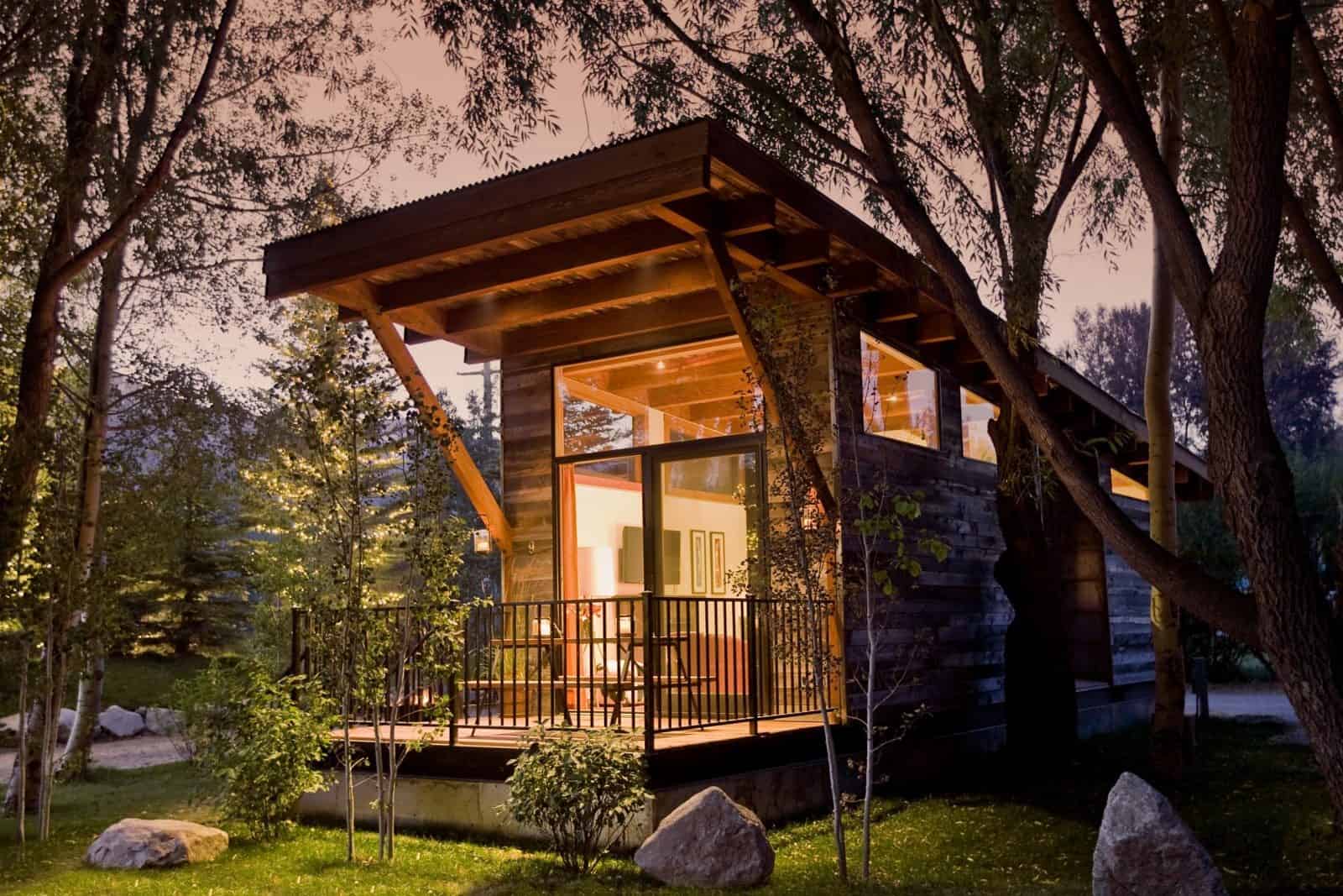 The Wedge at dusk under tall trees, with its angled roof, warm light glowing through tall windows, and a railed front deck