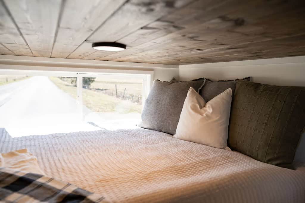 Inside the Wind River Pingora sleeping nook looking along the queen bed toward a long horizontal window at pillow height framing a rural road and field, with a stained pine plank ceiling and flush disc light overhead