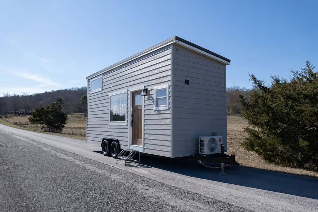 Three-quarter exterior view of the grey Wind River Pingora tiny house on wheels beside a rural road, showing the low shed roof slope, the wood entry door with metal steps, and a white mini-split condenser mounted at the rear corner
