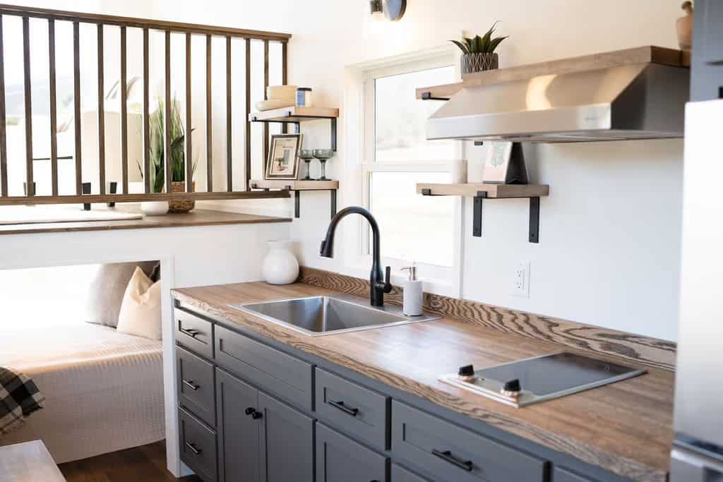 The Wind River Pingora galley kitchen: grey shaker base cabinets with black hardware, a figured butcher block counter, stainless single-basin sink beneath a window with a black gooseneck faucet, wood floating shelves on black brackets, stainless range hood, and the loft railing and bed nook in the background