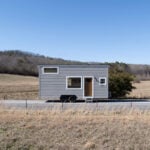 Grey Pingora tiny house on a dual-axle trailer on a rural road: horizontal grey lap siding, white-trim windows, wood door with metal steps down, dry grass and a wire fence in the foreground