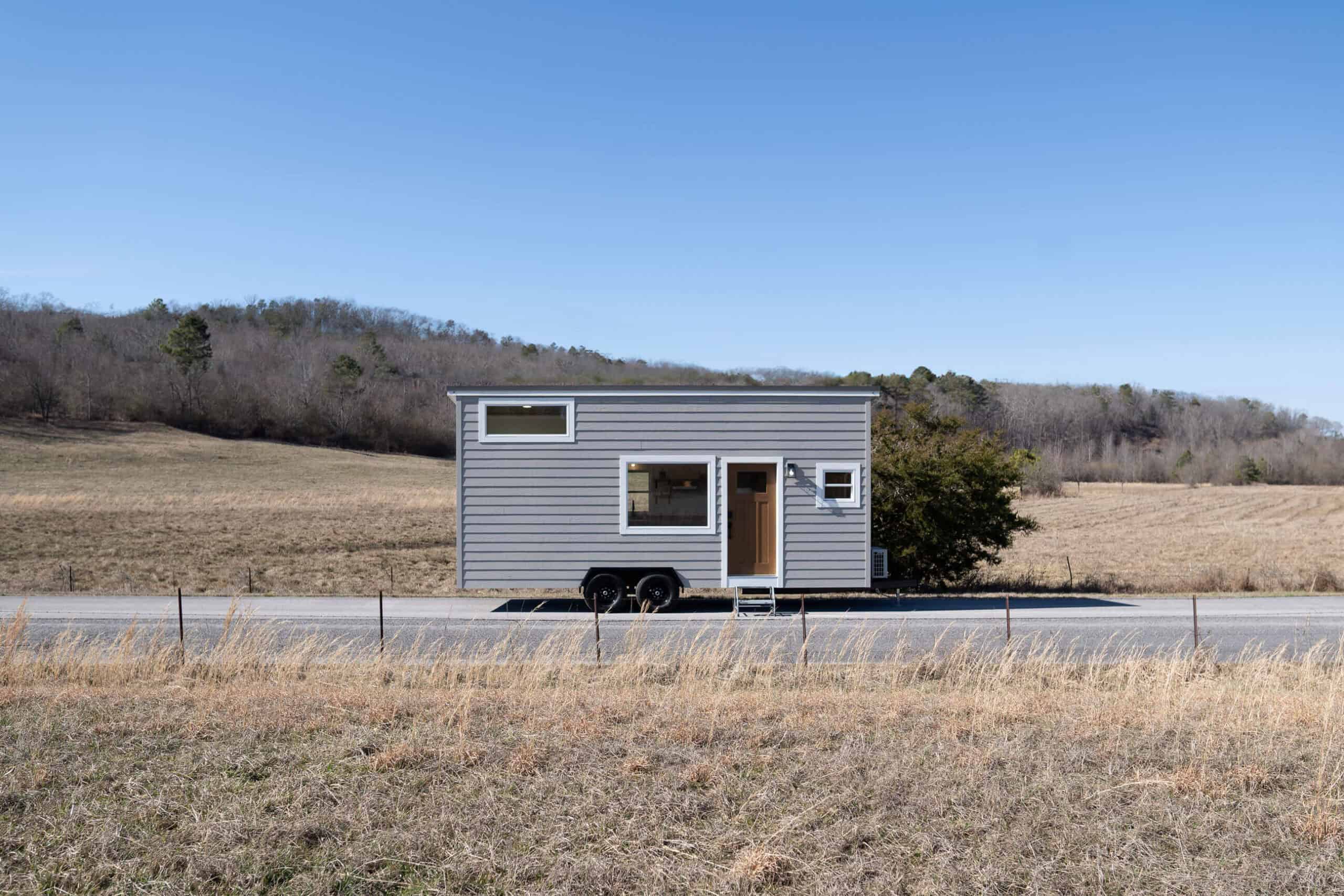 Grey Pingora tiny house on a dual-axle trailer on a rural road: horizontal grey lap siding, white-trim windows, wood door with metal steps down, dry grass and a wire fence in the foreground