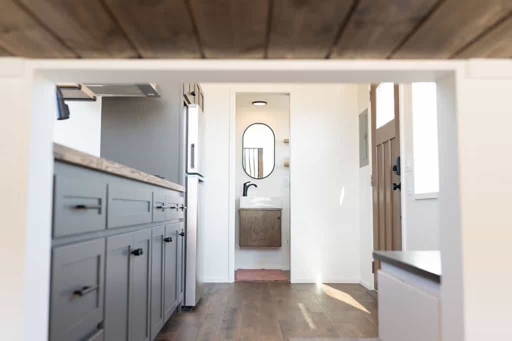 View down the length of the Wind River Pingora from the loft: grey shaker kitchen cabinets and stainless fridge on the left, an open bathroom doorway ahead showing a floating wood vanity with a rectangular sink and round black mirror, the wood entry door at right, and a stained plank ceiling overhead