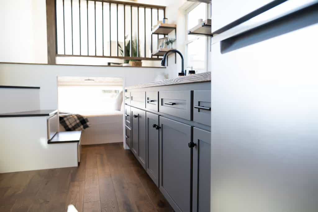Low-angle interior view of the Wind River Pingora showing dark-stained wood plank flooring, a run of grey shaker cabinets and drawers with black hardware on the right, open stair treads with storage boxes below on the left, and the bed nook and loft railing beyond