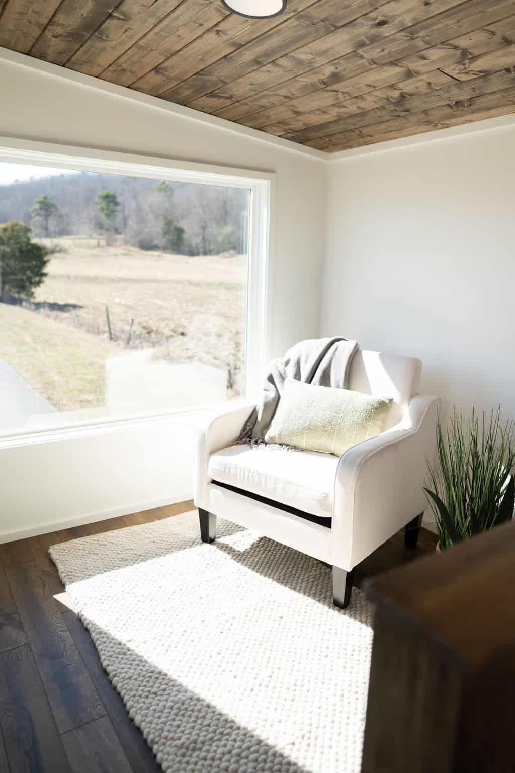 The Wind River Pingora lofted living area: a white upholstered armchair with a throw and pillow on a chunky cream rug, beside a large low picture window framing a winter field and treeline, under a rough-sawn stained pine plank ceiling with a flush disc light