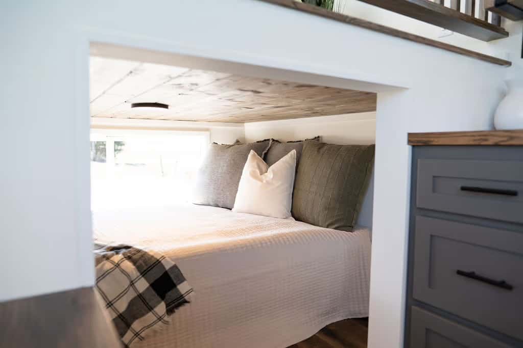 The main-floor sleeping nook of the Wind River Pingora framed as a built-in opening beneath the lofted living platform, with a queen bed made up in white bedding, layered grey and olive pillows, and a plaid throw, beside grey kitchen drawers