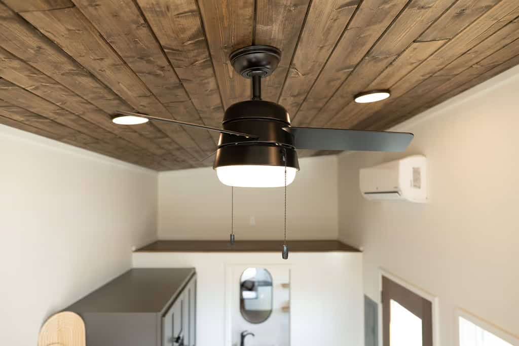 Looking up in the Wind River Pingora at a black ceiling fan with light hung from a rough-sawn stained pine plank ceiling, a white LG mini-split head mounted high on the end wall, and recessed disc lights, with the bathroom mirror and entry door visible below