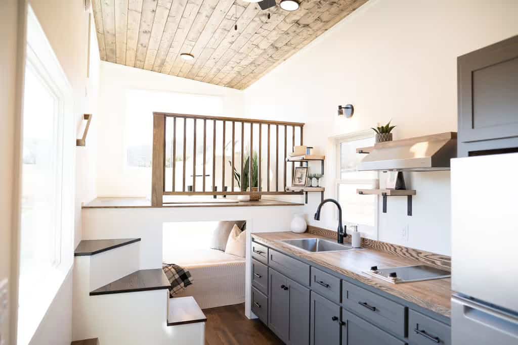 Interior of the Wind River Pingora looking down the galley kitchen with grey shaker cabinets and butcher block counters, with open stair treads on the left climbing to a lofted living platform behind a wood spindle railing, and the main-floor bed visible in a nook beneath the platform