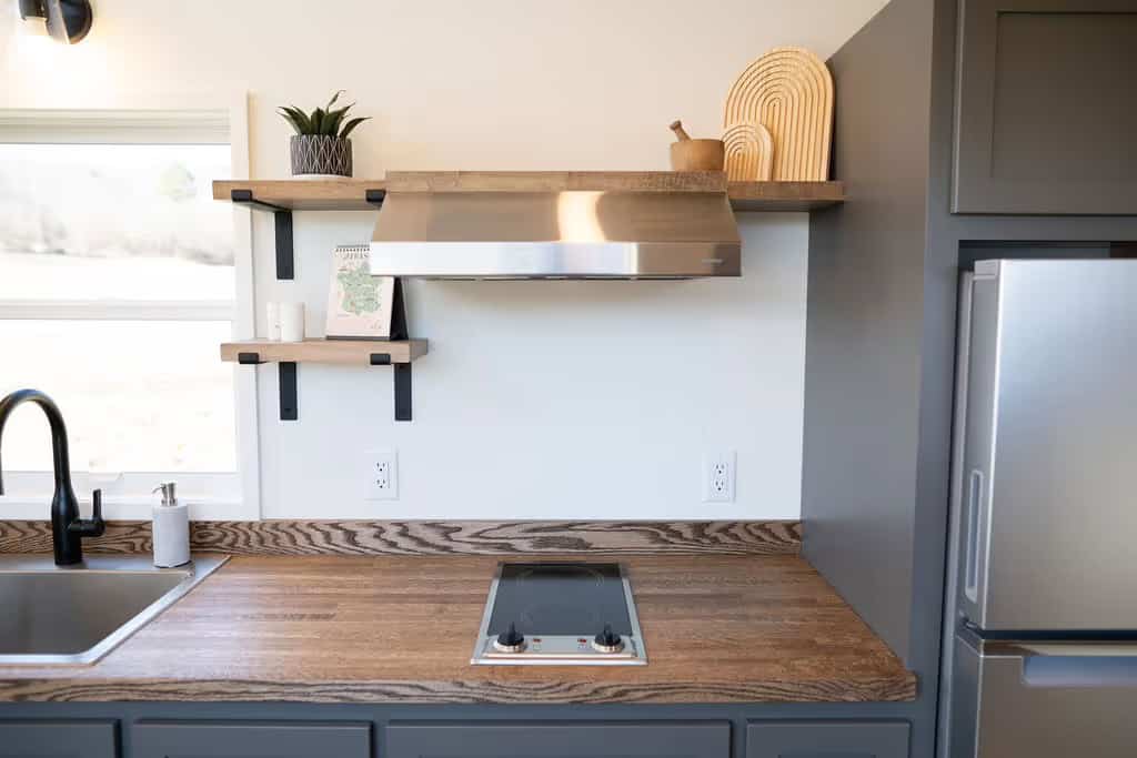 Close view of the Wind River Pingora cooking area: a two-burner electric cooktop set flush into the butcher block counter beneath a full-width stainless steel range hood, with two wood floating shelves on black brackets holding a plant and cutting boards, and a stainless fridge at right