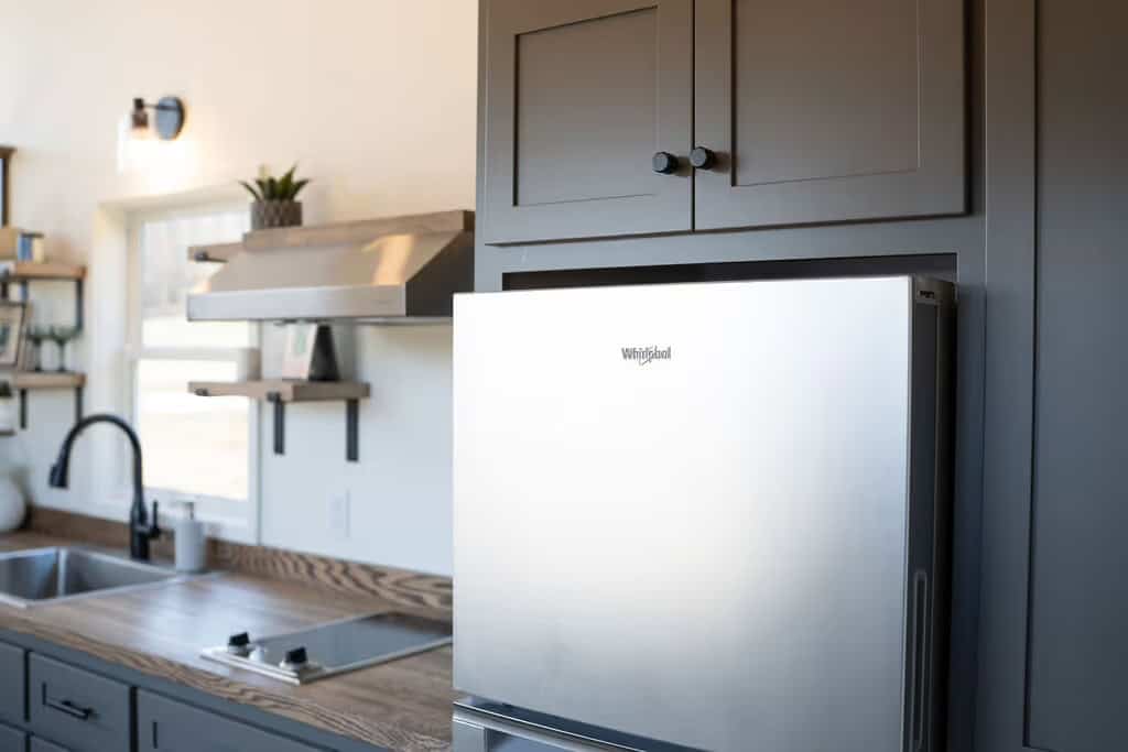 A stainless Whirlpool three-quarter-size refrigerator built into a grey cabinet surround in the Wind River Pingora kitchen, with a grey shaker upper cabinet above it and the butcher block counter, cooktop and sink visible beyond