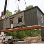 A crane lifts a modular student cottage over a wooded campus construction site, with another cottage already set on its foundation