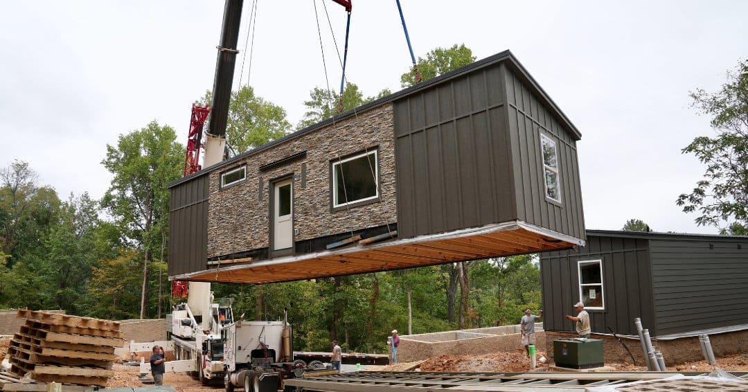 A crane lifts a modular student cottage over a wooded campus construction site, with another cottage already set on its foundation