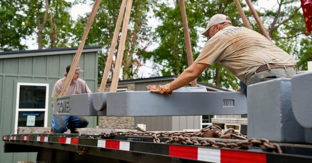 Two workers on a flatbed trailer with crane counterweights, a sage-green modular cottage numbered 19 in the trees behind them