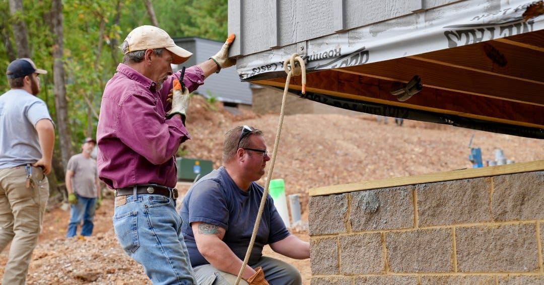 Two workers guide a gray board-and-batten modular cottage onto a concrete-block foundation, wooden floor joists visible underneath