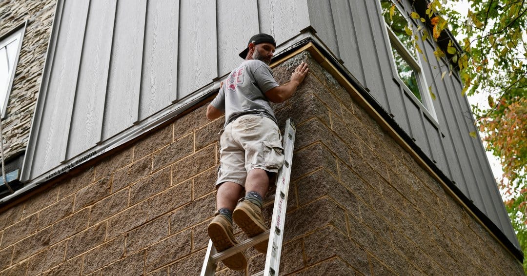 A worker on a ladder finishes the corner where gray board-and-batten siding meets the stone foundation of a modular cottage
