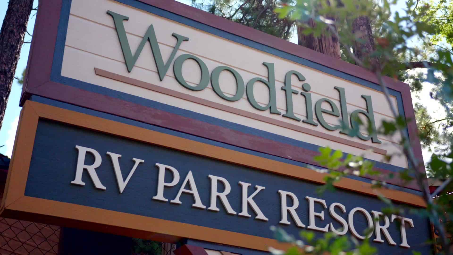 Entrance sign reading Woodfield RV PARK RESORT, with pines behind it