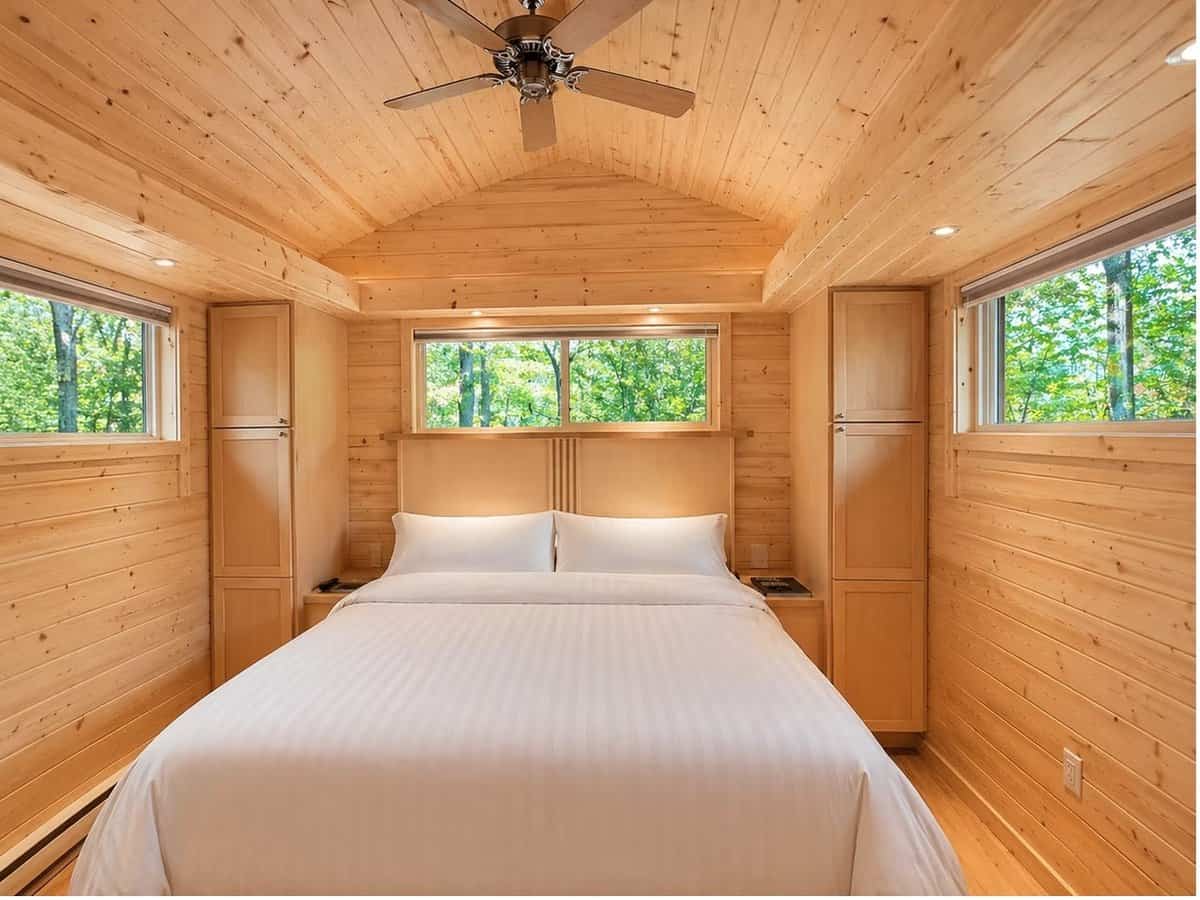 Walk-around king bed under a vaulted pine ceiling flanked by built-in nightstands and storage towers with forest views at the Woodland Lot 12