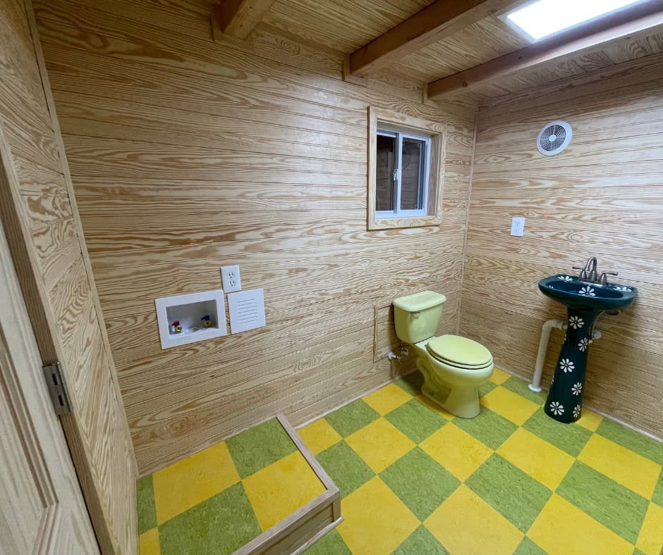 Bathroom with natural wood walls, a pale yellow toilet, the painted pedestal sink, washer and dryer hookups, a window, and a checkerboard floor