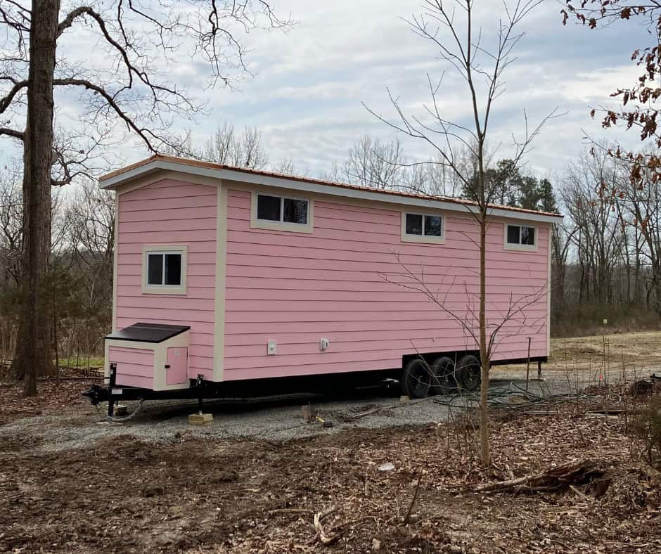 Pink tiny house on wheels seen from the front end, with a small pink exterior storage box and high windows along the side