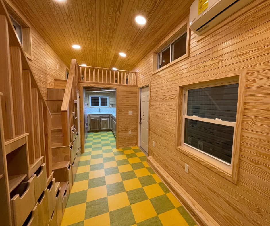 Main room with natural wood walls and ceiling, storage stairs on the left, a large window and entry door on the right, and the kitchen beyond under a loft rail