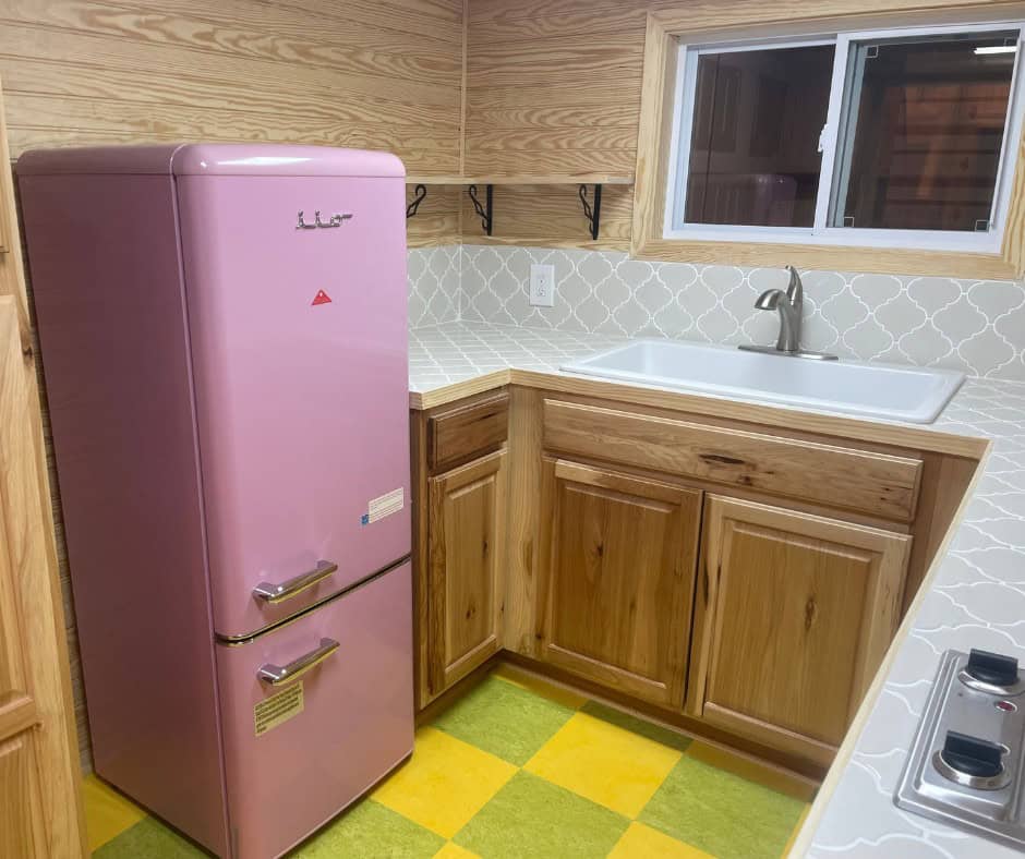 Pink retro-style refrigerator beside hickory base cabinets and a white sink under a window