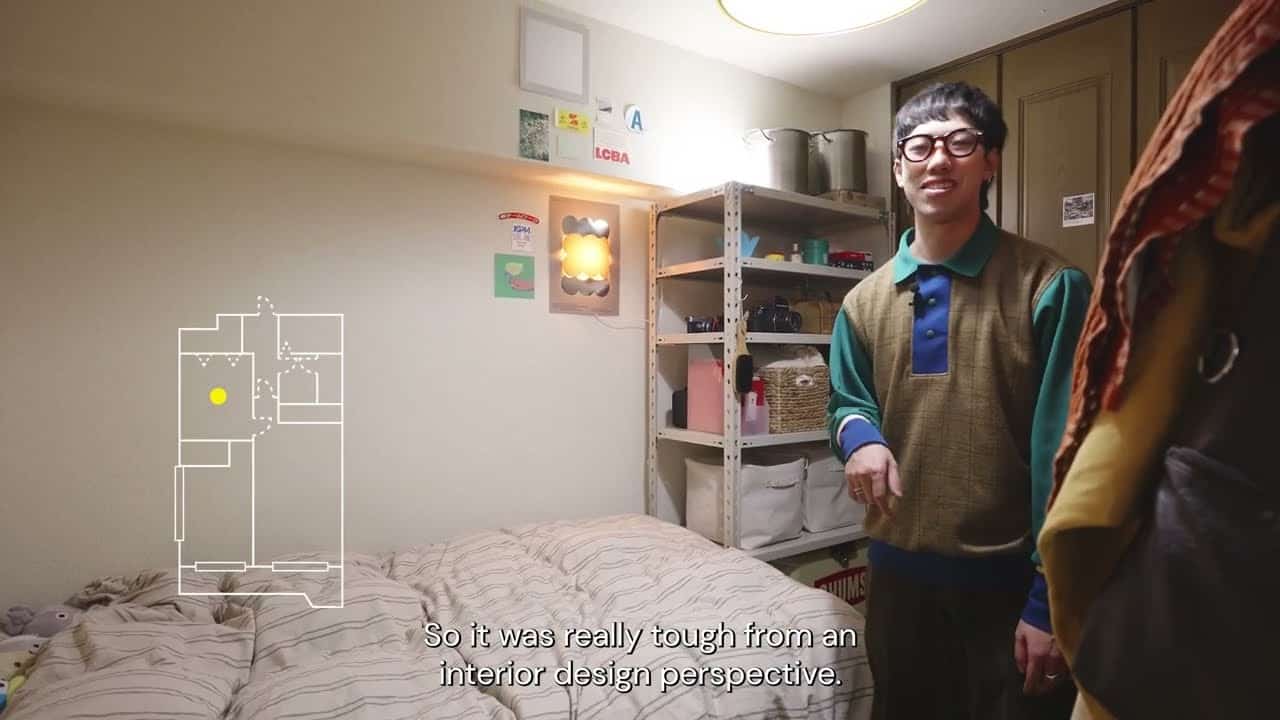 Ryu standing beside the bed in the Yoshinani bedroom, open steel shelving packed with pots and baskets