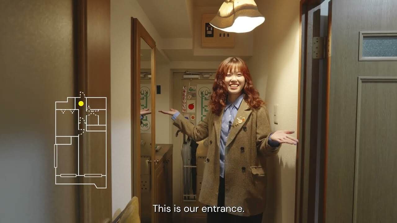 Kamado standing in the narrow Yoshinani House entrance hallway, smiling, with a floor-plan overlay and the subtitle This is our entrance