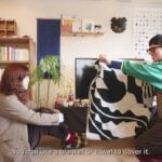Ryu kneeling in the Yoshinani living room, holding a black-and-white patterned blanket up to cover the television, Kamado seated on the floor