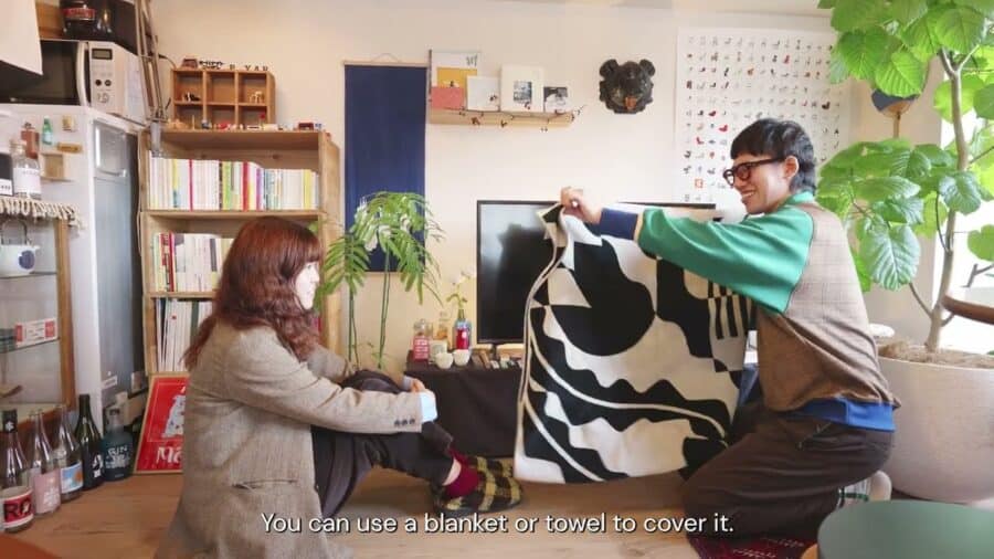 Ryu kneeling in the Yoshinani living room, holding a black-and-white patterned blanket up to cover the television, Kamado seated on the floor